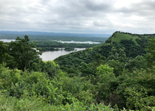 Lush green hills and a river valley under a cloudy sky, showcasing a scenic landscape.