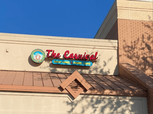 Sign for "The Carnival" featuring snow cones and funnel cakes, mounted on a building under a clear blue sky.