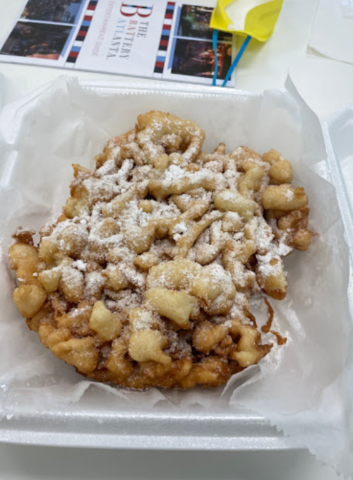 A freshly made funnel cake dusted with powdered sugar, served in a white takeout container.