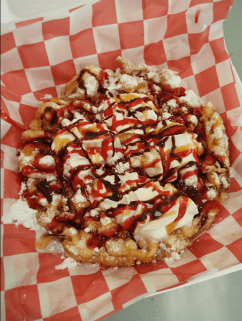 A funnel cake topped with whipped cream, chocolate syrup, and strawberry sauce, served in a red and white checkered paper.