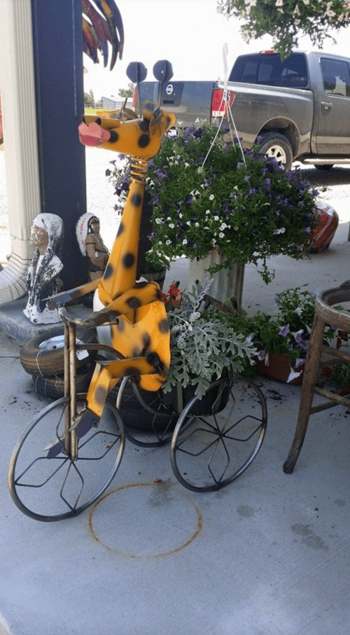 A whimsical metal giraffe on a bicycle surrounded by colorful flowers and decorative items.