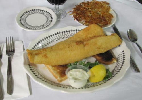 A plate with a large fried fish fillet, tartar sauce, lemon, and hash browns, served with a glass of dark beverage.