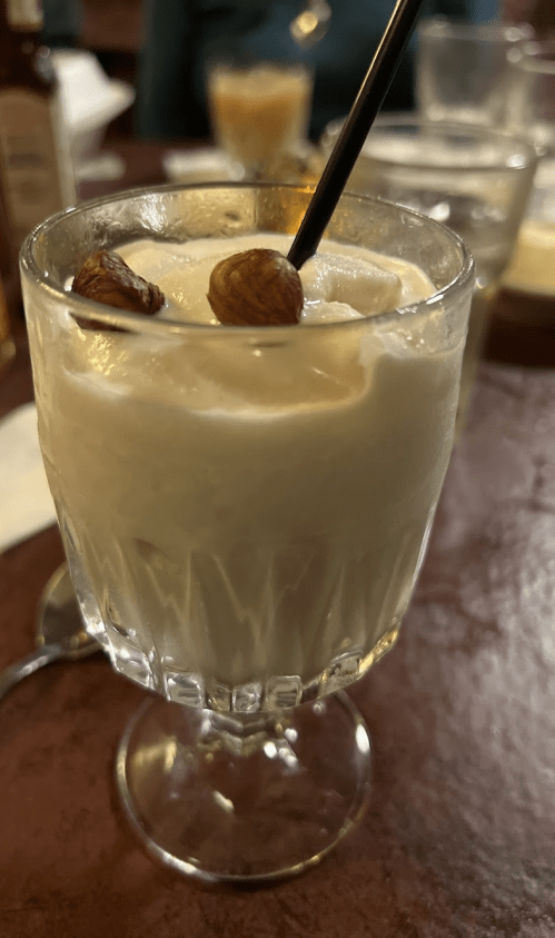 A creamy dessert drink in a glass, topped with nuts and a straw, sitting on a wooden table.