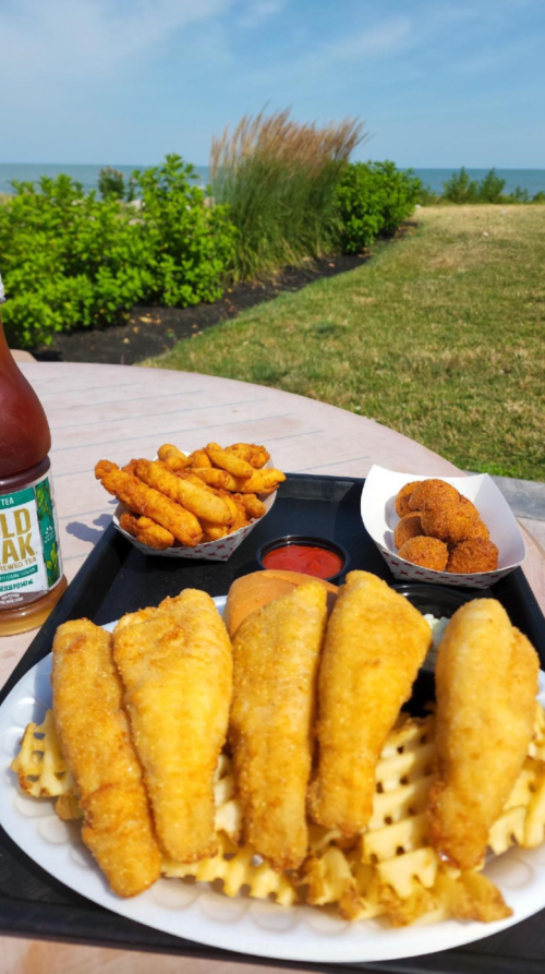 A tray of golden fried fish fillets on waffle fries, with hush puppies and a side of dipping sauce, set outdoors by the water.