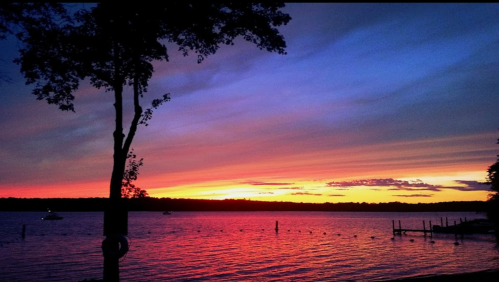 A vibrant sunset over a lake, with colorful skies and silhouettes of trees and boats in the distance.