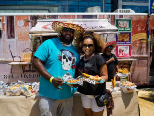 A man in a sombrero and a woman hold food in front of a food truck, smiling at a sunny outdoor event.