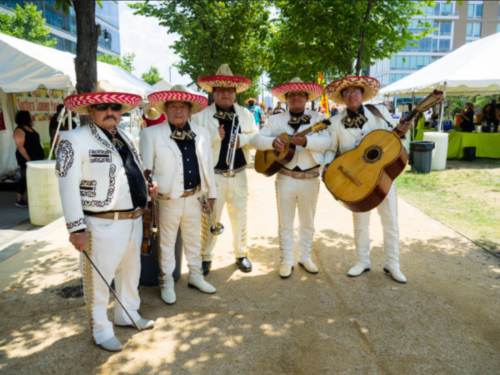 A mariachi band in traditional attire poses outdoors, surrounded by trees and festival tents.