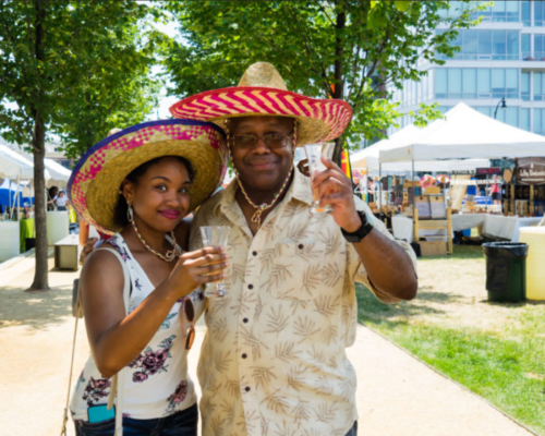 A man and woman wearing colorful sombreros hold drinks, smiling at a sunny outdoor market with tents in the background.