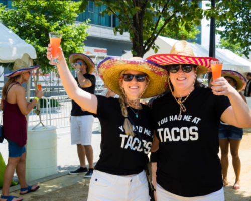 Two women in sombreros hold drinks, wearing matching shirts that say "You had me at Tacos," smiling at a festival.