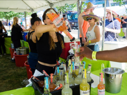 A bartender pours a colorful drink at a festival booth, with people enjoying the event in the background.