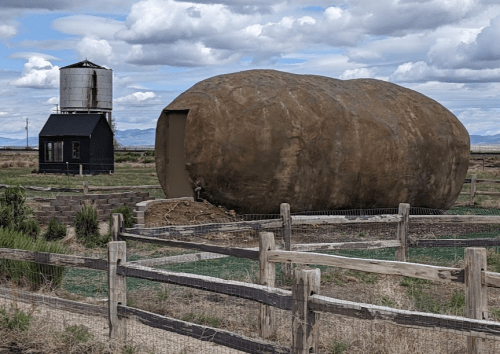 A large, potato-shaped structure sits in a field near a black building and a water tower under a cloudy sky.