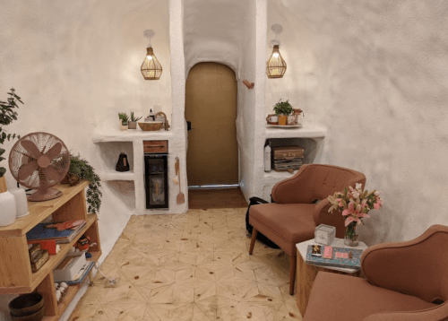 Cozy interior with white walls, two chairs, a wooden shelf, plants, and warm lighting, creating a welcoming atmosphere.