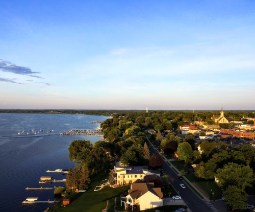 Aerial view of a lakeside town with boats, trees, and buildings under a clear blue sky.