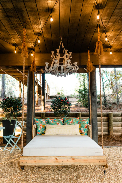 A cozy outdoor swing bed with colorful cushions, surrounded by string lights and greenery under a wooden structure.