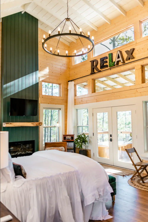 Cozy bedroom with a large window, chandelier, and "RELAX" sign, featuring a white bed and wooden accents.