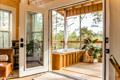 A cozy interior with an open door leading to a deck featuring a hot tub and plants, surrounded by trees.