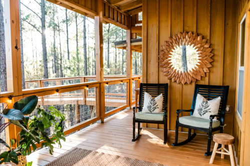 A cozy porch with two rocking chairs, a decorative mirror, and greenery, surrounded by tall trees.