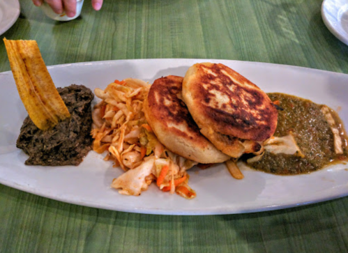 A plate featuring two arepas, plantains, coleslaw, black beans, and green sauce on a green table.
