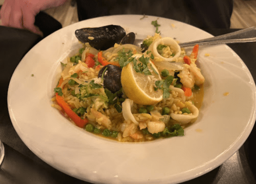 A bowl of seafood paella with shrimp, squid, mussels, and vegetables, garnished with lemon and herbs.