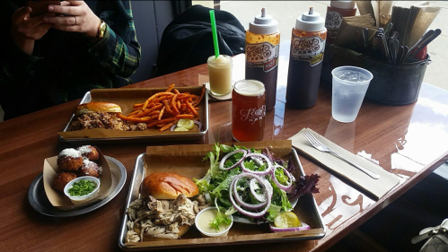A table with various dishes: pulled pork sandwich, sweet potato fries, fried balls, salad, and drinks.