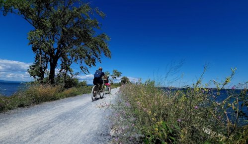 Two cyclists ride along a gravel path by a lake, surrounded by trees and wildflowers under a clear blue sky.
