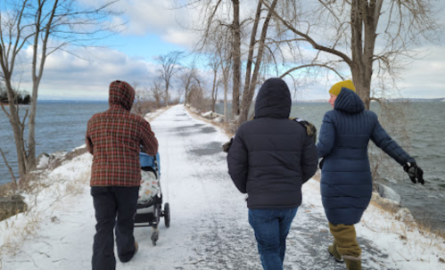 Three people walk along a snowy path by the water, with trees lining the way and a stroller in tow.