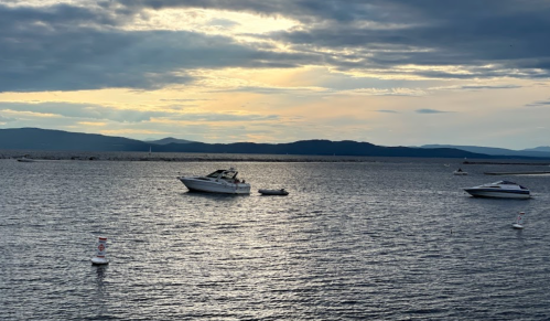 Boats floating on calm water under a cloudy sky at sunset, with distant hills in the background.