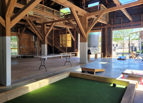 Interior of a rustic barn with wooden beams, a bocce ball court, and tables set up for an event.