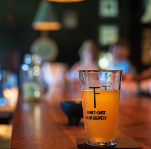 A glass of beer on a wooden bar, with a logo for Towerhouse Farm Brewery, and blurred figures in the background.
