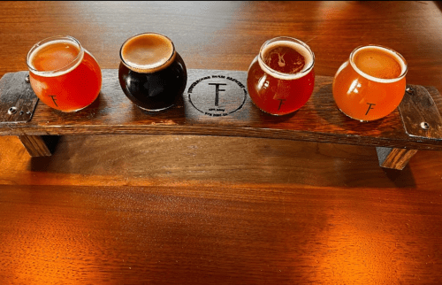 A wooden flight board holds four small beer glasses, each with different colored brews, on a wooden table.