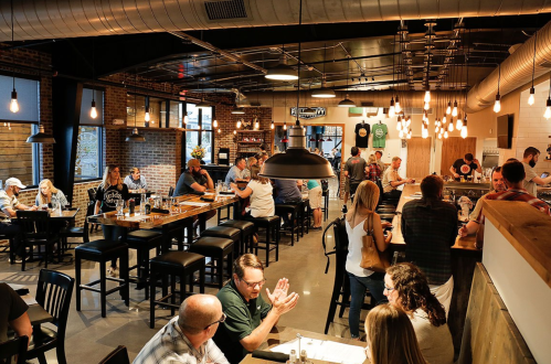 A lively restaurant interior with patrons dining, chatting, and a bar area, featuring modern decor and warm lighting.