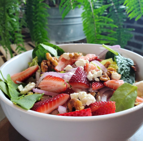 A fresh salad in a white bowl, featuring spinach, strawberries, walnuts, red onion, and feta cheese, with greenery in the background.
