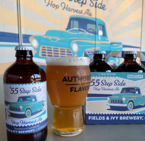 A bottle and a glass of '55 Step Side Hop Harvest Ale next to a six-pack, with a vintage truck backdrop.