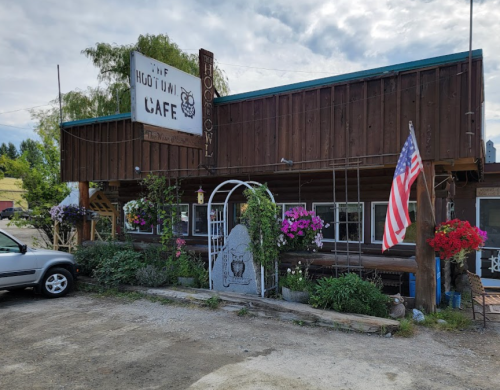 A rustic café with a wooden exterior, flower baskets, and an American flag outside, set in a rural area.