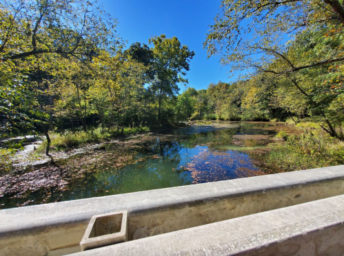 A serene pond surrounded by trees, reflecting blue sky and autumn foliage, viewed from a stone railing.