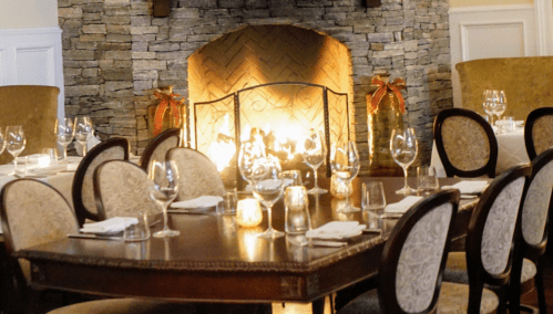 Cozy restaurant interior with a stone fireplace, elegant dining table, and set tables ready for guests.