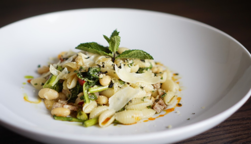 A bowl of pasta with greens, white beans, and garnished with mint and cheese shavings on a wooden table.