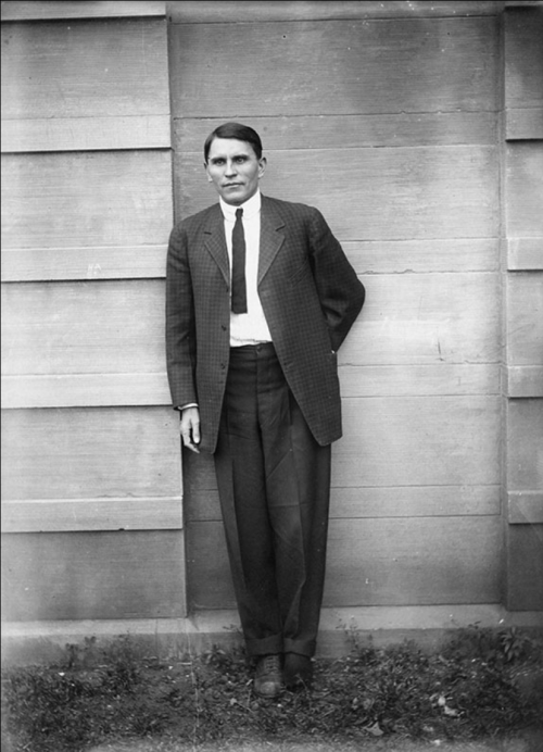 A man in a suit stands against a wooden wall, hands behind his back, with a serious expression.