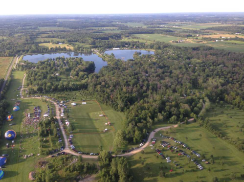Aerial view of a lush landscape with lakes, forests, and a gathering area with tents and parked cars.
