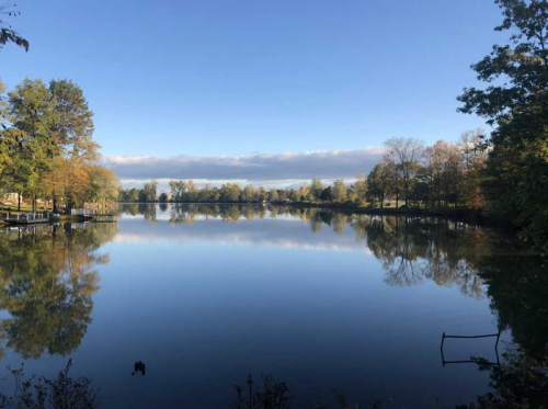 A serene lake reflecting trees and a clear blue sky, with autumn colors along the shore.