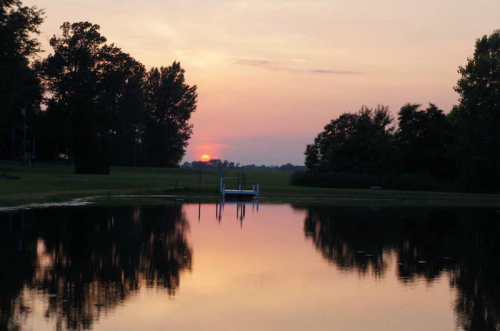 A serene sunset over a calm pond, reflecting trees and a dock in the water.