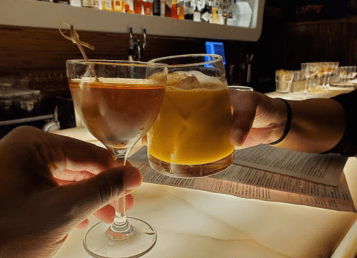 Two hands clink glasses at a bar, one holding a cocktail and the other a drink with ice. Soft lighting in the background.