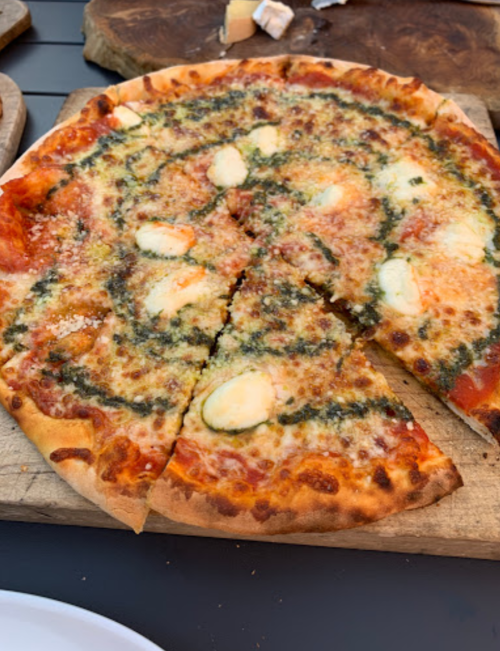 A freshly baked pizza with melted cheese, pesto, and sliced pieces on a wooden board.