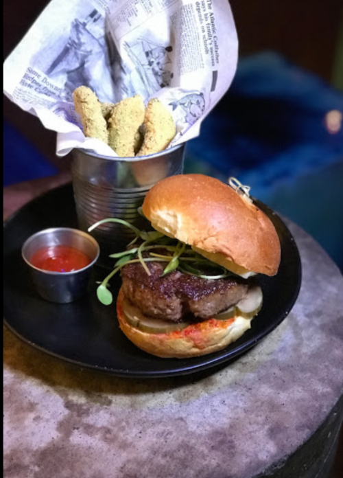 A gourmet burger with greens on a bun, served with a side of fried okra in a cup and a small bowl of sauce.