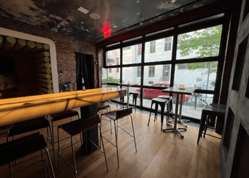 Interior of a modern bar with a wooden floor, large windows, and a glowing bar counter. Outdoor view visible.