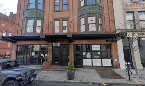 A brick building with large windows and a storefront, featuring a parked Jeep in front and street parking meters.