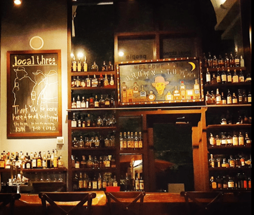 A cozy bar interior with shelves lined with various bottles of liquor and a welcoming sign on the wall.