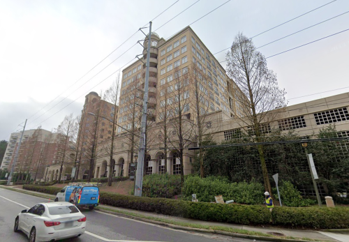 A street view of a multi-story building with trees and a parked van in the foreground, along a busy road.