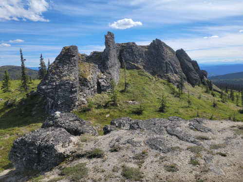 Rocky outcrop on a grassy hillside, surrounded by sparse trees under a blue sky with scattered clouds.