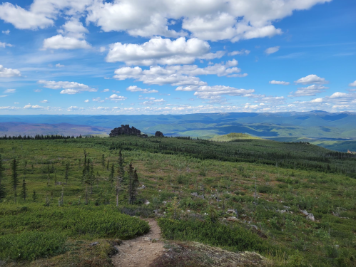 A scenic view of rolling green hills under a blue sky with fluffy clouds, featuring a rocky outcrop in the distance.